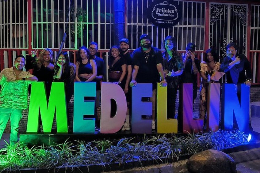 Un grupo de personas posando frente a un letrero grande iluminado con la palabra 'MEDELLUN' y un restaurante llamado 'Frijoles Banocos' al fondo. 5