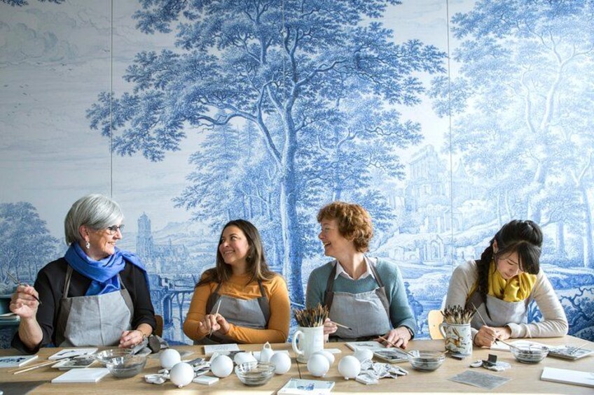 Cuatro personas sentadas en una mesa con cerámica y material de pintura, frente a un mural grande azul y blanco. 1