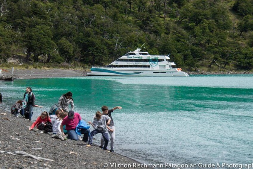 A group of people on a rocky shore, a large boat in the water, and a forested hill in the background. 2
