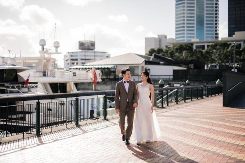A couple walking on a waterfront promenade with boats and buildings in the background. 1