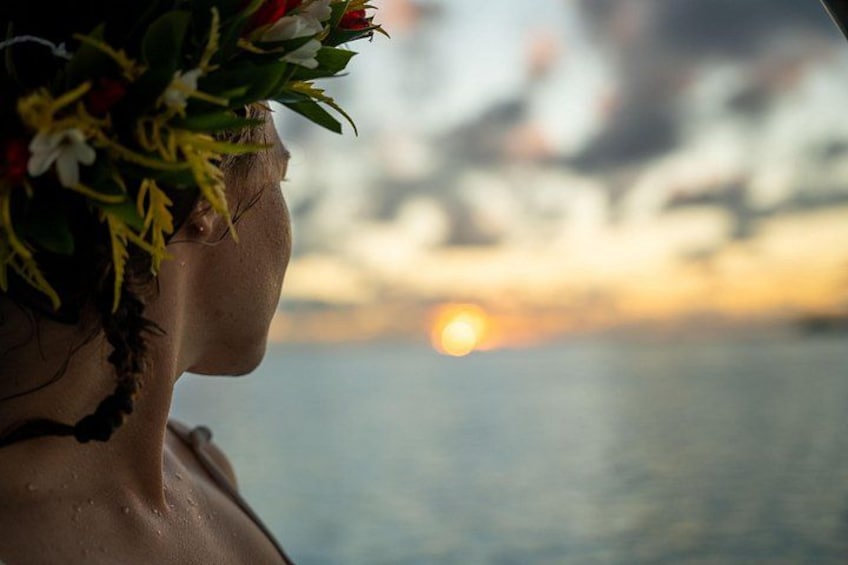 A person with a floral crown, facing a sunset over water. 5