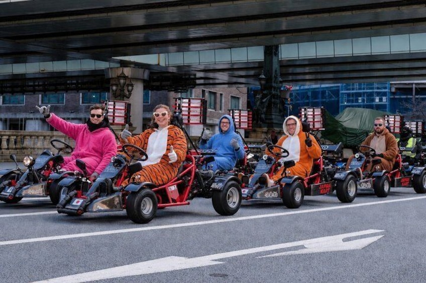 Personas en karts sobre una carretera, con edificios al fondo. 1