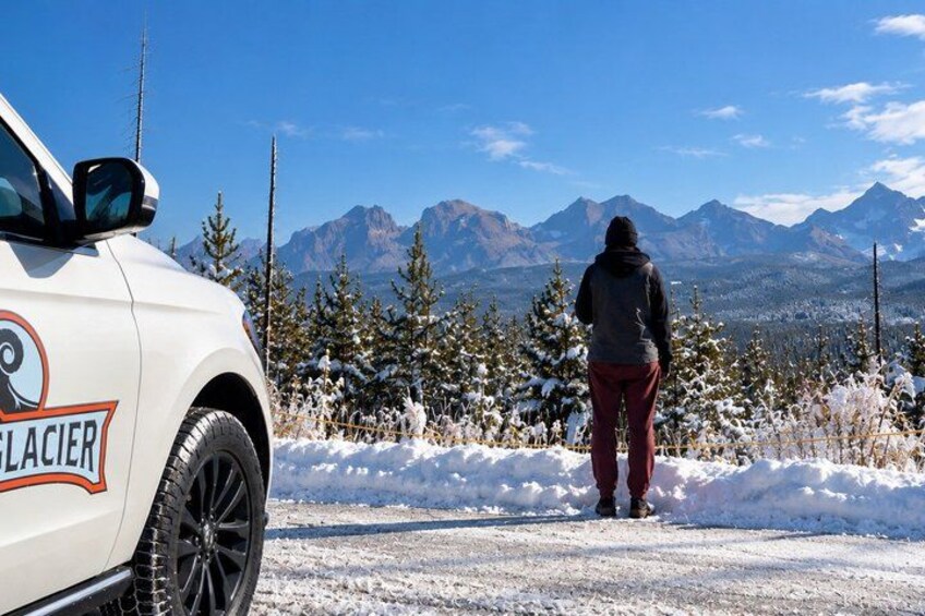 A person standing in a snowy landscape with mountains in the background and a vehicle labeled 'GLACIER' in the foreground. 1