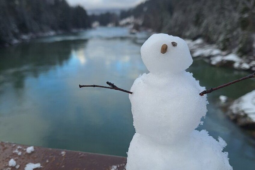 A snowman with sticks for arms, a river in the background, and snow-covered trees. 2