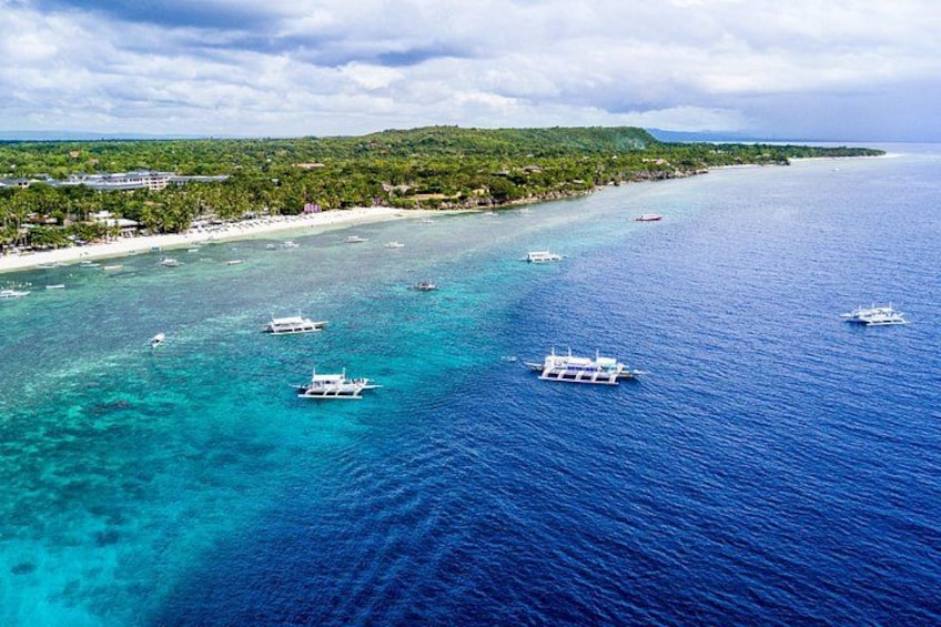 A coastal area with clear blue waters, several boats, and a beach with buildings in the background. 3