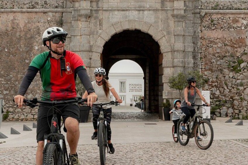 Des personnes à vélo circulent près d’une arche en pierre, avec un panneau visible à l’arrière-plan. 1