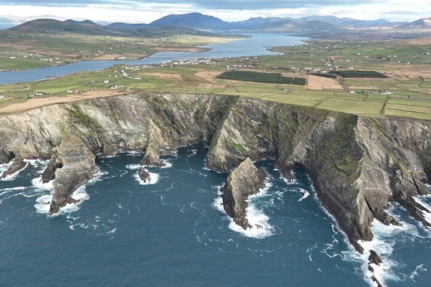 A coastal cliff with waves crashing against it, a distant body of water, and a landscape with fields and buildings. 3