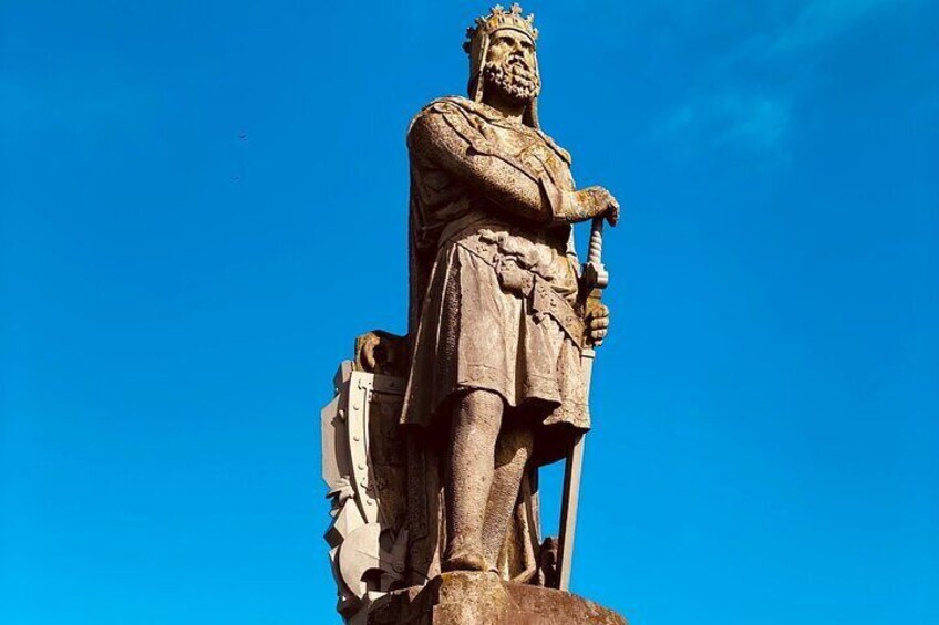 A stone statue of a king holding a scepter and a sword, standing on a pedestal against a clear blue sky. 5