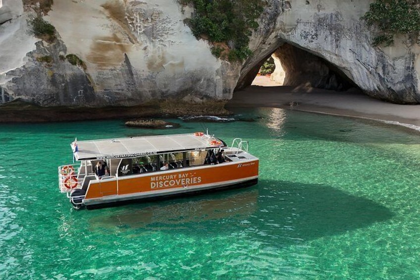 Un bote llamado 'Mercury Bay Discoveries' sobre aguas turquesas, con una formación rocosa tipo cueva al fondo. 5