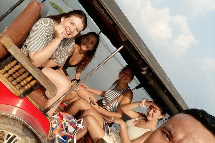 A group of people sitting on a red vehicle with a wooden bench, a steering wheel, and a steering column. 5