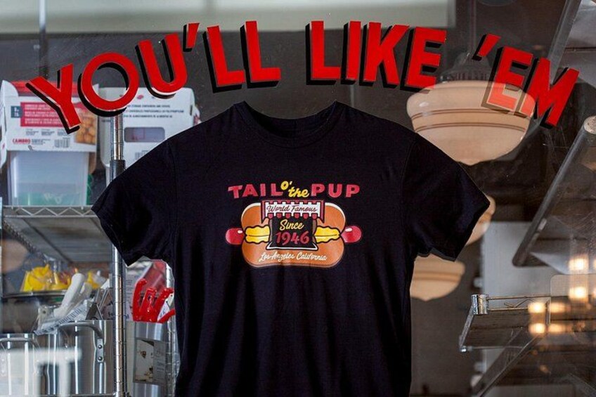 Una camiseta negra con un gráfico y texto, exhibida en una tienda con estanterías y productos de fondo. 3