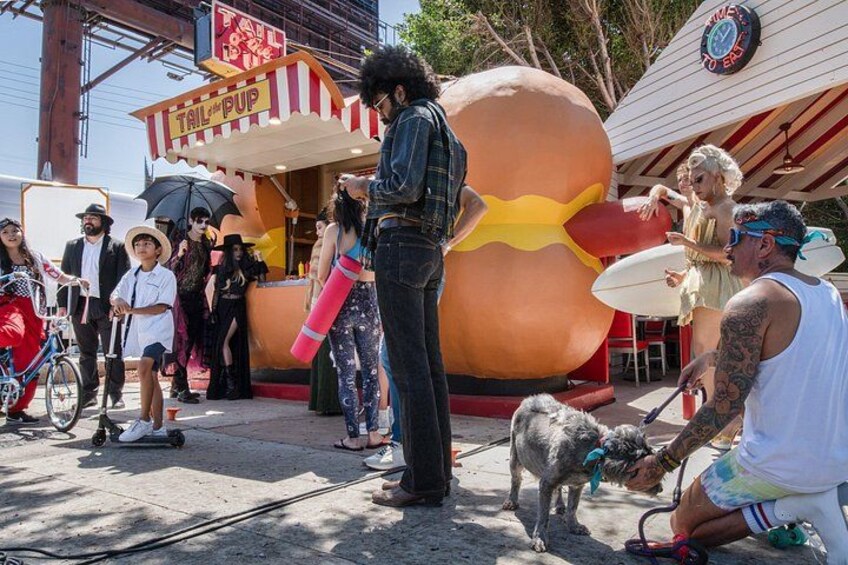 Escena callejera con una gran escultura de hot dog, un puesto de comida y gente caminando. 2
