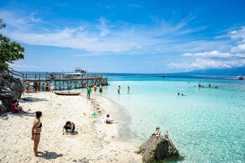 Een strand met helder turquoise water, een houten steiger en mensen die zwemmen en ontspannen aan de kust. 2