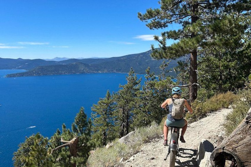 A person riding a bicycle on a dirt trail with a mountainous landscape and a body of water in the background. 2