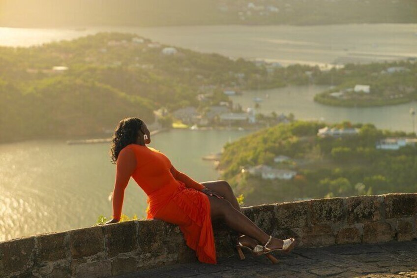 A person in an orange dress sitting on a stone wall overlooking a body of water with buildings and greenery in the background. 2