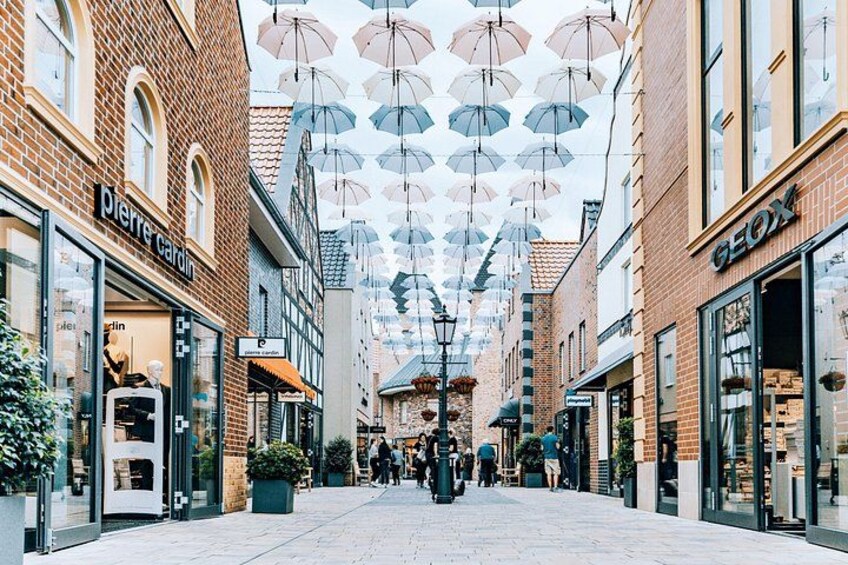 A pedestrian street lined with shops, featuring a row of hanging umbrellas and potted plants. 1
