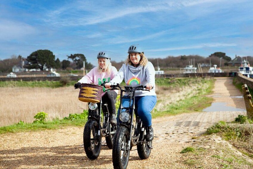 Two people riding bicycles on a rural path with boats in the background. 4