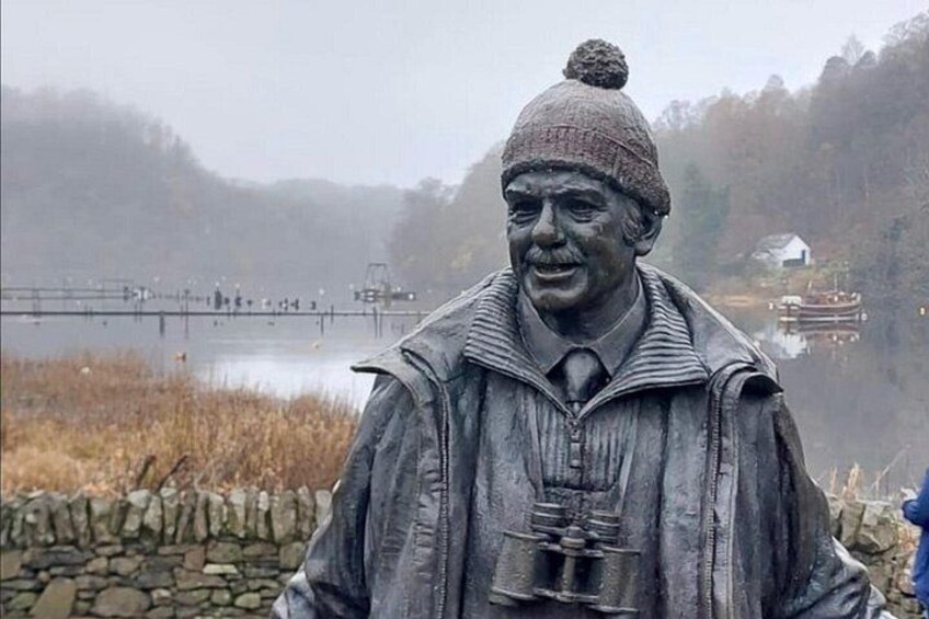 Una estatua de un hombre con gorro, en un entorno natural con una masa de agua y un muro de piedra al fondo. 5