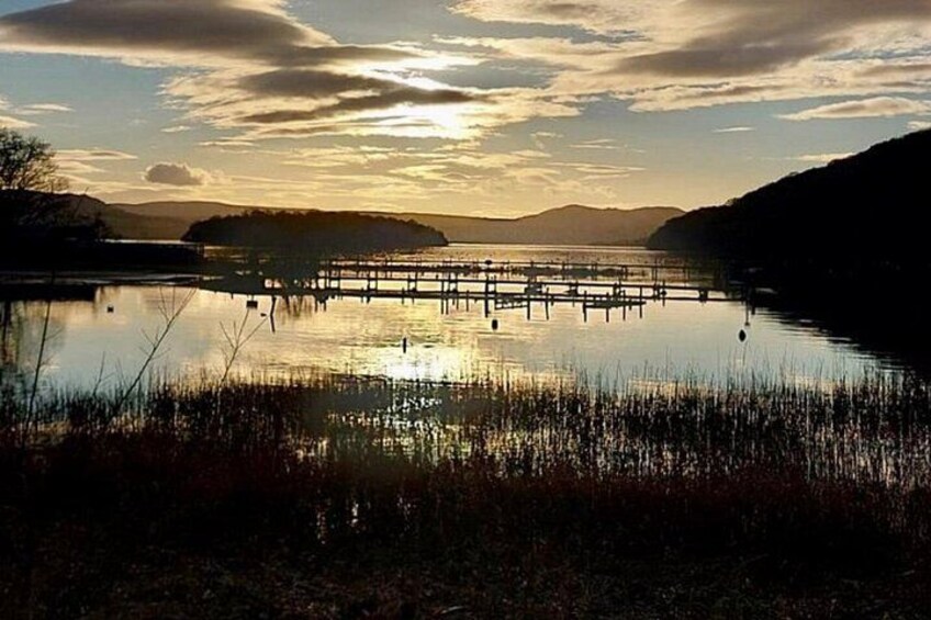 Un lago tranquilo con un muelle de madera que se adentra en el agua, rodeado de orillas cubiertas de hierba y colinas distantes bajo un cielo parcialmente nublado. 4