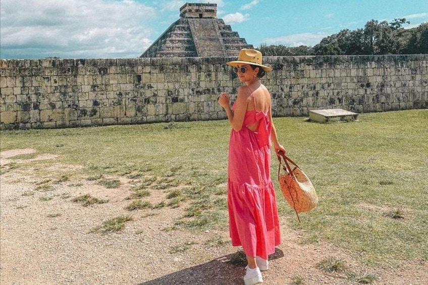 Eine Person in einem pinkfarbenen Kleid und gelbem Hut steht vor einer Steinmauer, im Hintergrund ist eine Pyramide zu sehen. 1