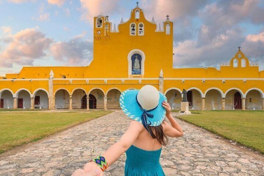 A woman in a blue dress and white hat standing in front of a yellow church with arches. 3