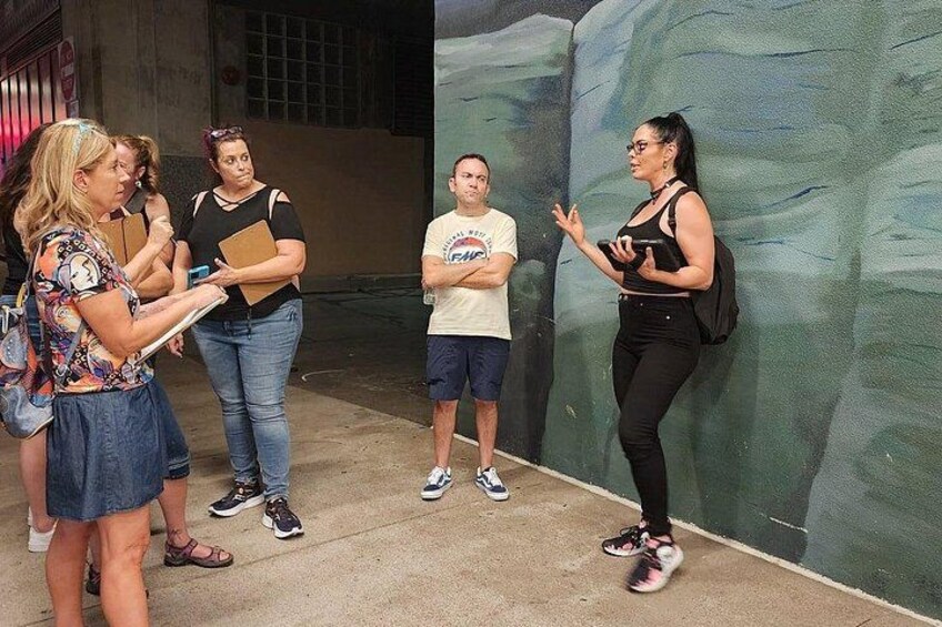 A group of people standing outdoors, some holding papers, in front of a mural. 2