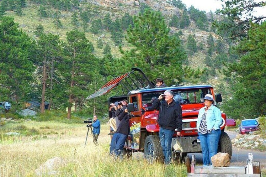 A group of people around a red vehicle with an open roof, in a grassy area with trees and hills in the background. 1