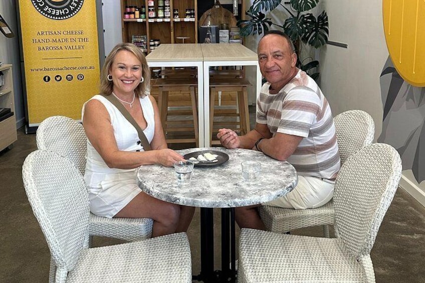 Two people sitting at a round table with a plate of food, in a room with wicker chairs and a yellow wall. 1