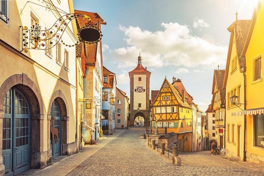 A cobblestone street lined with colorful half-timbered houses, a clock tower, and a clear sky. 5