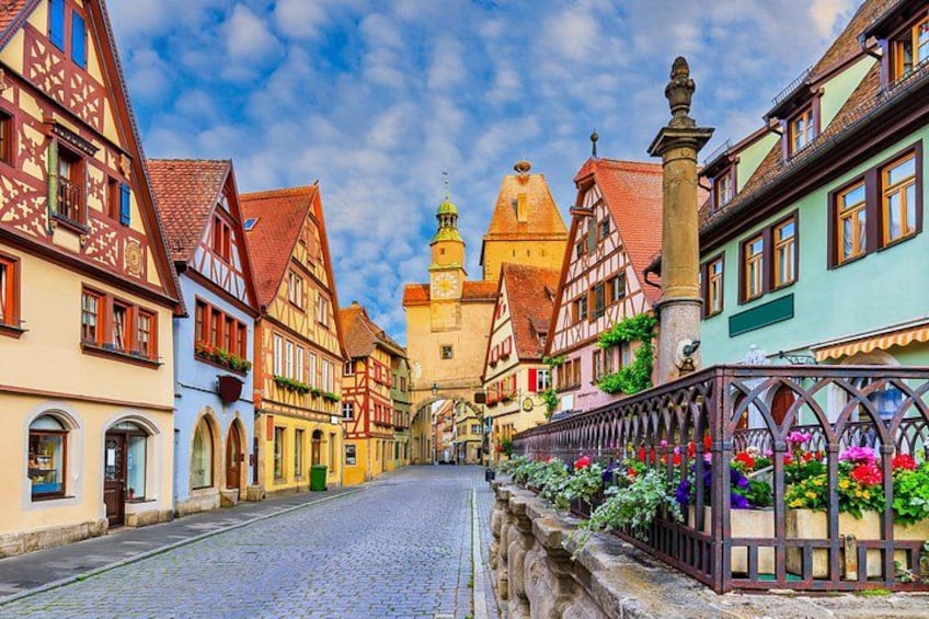 A cobblestone street lined with traditional half-timbered houses, a church steeple, and a flower-adorned fence. 1