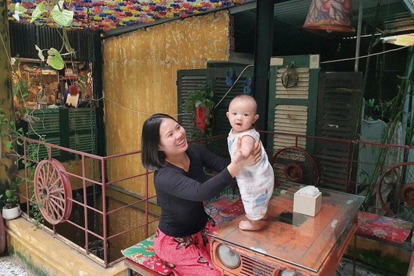 A woman holding a baby in a rustic outdoor setting with a vintage radio, a wheelbarrow, and a small shop in the background. 4