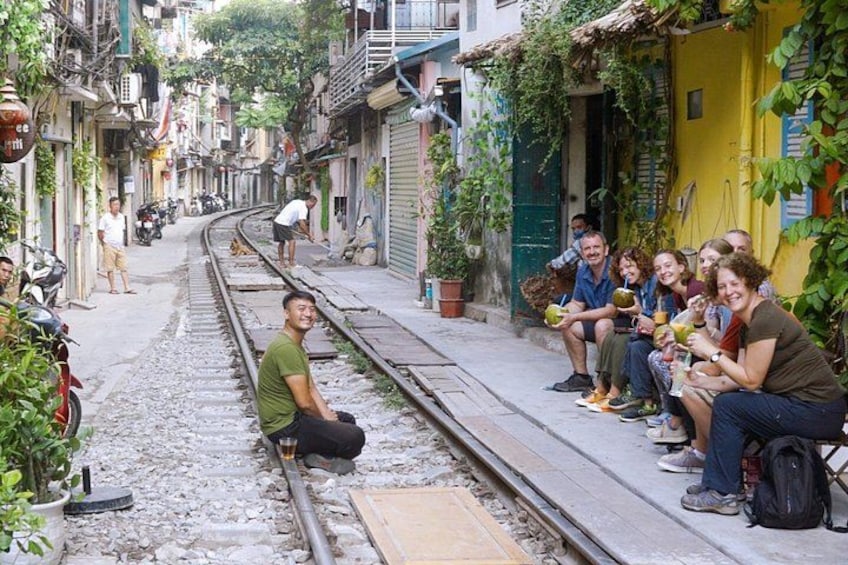 A narrow street with a railway track, people sitting on the sidewalk, and buildings with balconies and greenery. 2