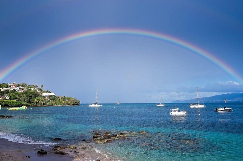 Eine Küstenlandschaft mit Regenbogen, klarem blauen Wasser, Booten und einem grünen Hügelhang. 3