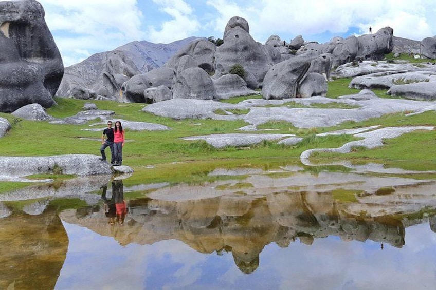 A couple standing on a rock by a reflective water body with large boulders in the background. 1