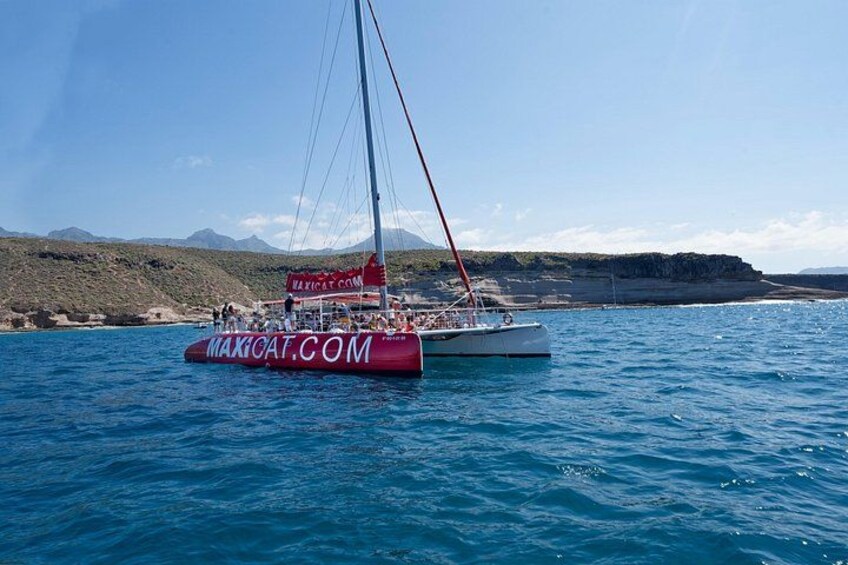 A sailboat with a red hull and a white superstructure, sailing on a calm sea with a mountainous coastline in the background. 4