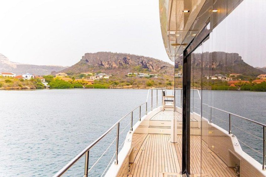 A boat deck with a view of a calm lake, distant rocky cliffs, and a few buildings on the shore. 5