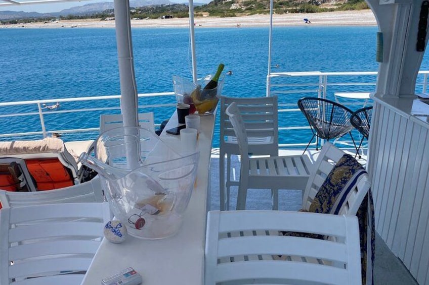 A boat deck with white chairs, a table with a bottle and glasses, and a view of the sea and beach. 2