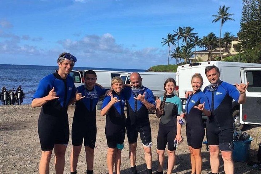 A group of people in wetsuits posing on a beach with palm trees and a vehicle in the background. 3