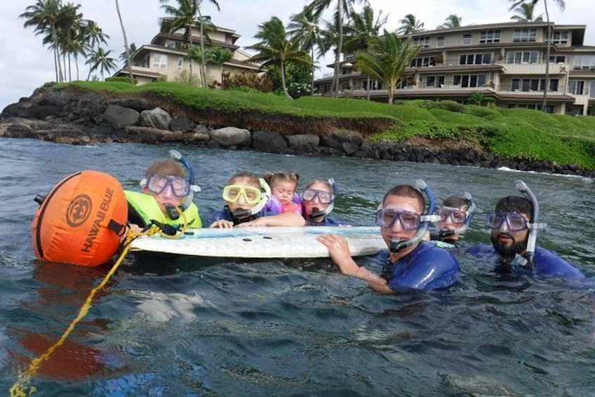 A group of people in blue wetsuits on a surfboard in the ocean with a Hawaiian Blue buoy and a building in the background. 1