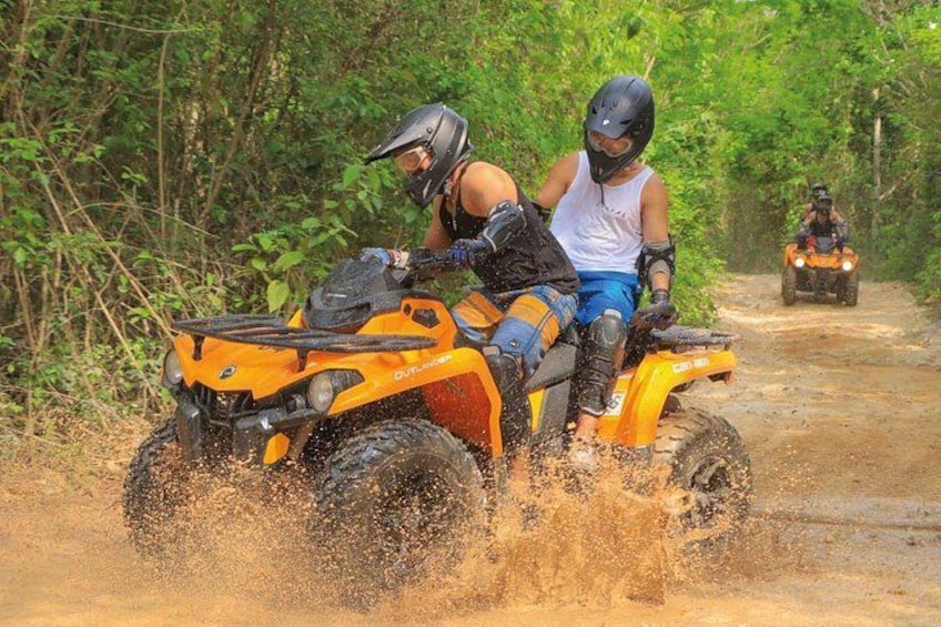 Two people riding an orange ATV on a muddy path surrounded by greenery. 4