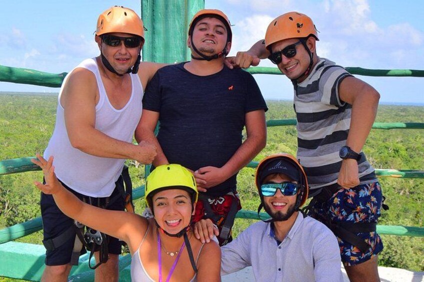 A group of people wearing helmets and harnesses, standing on a platform with a green railing, overlooking a landscape. 2
