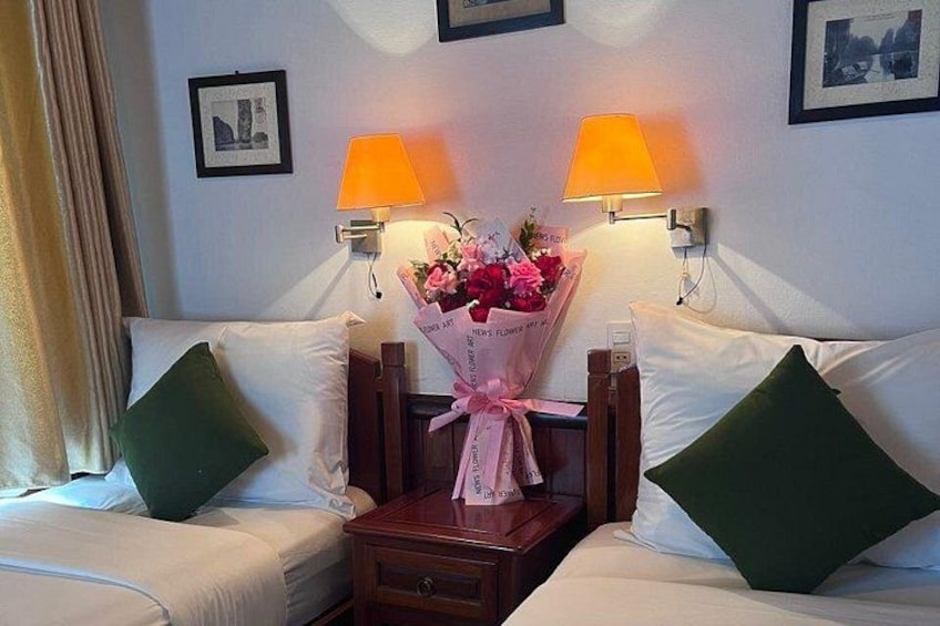 A hotel room with two beds, green pillows, a nightstand with a lamp, and a bouquet of flowers. 4