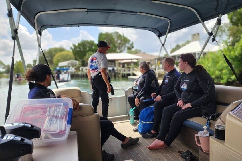 People on a boat, some wearing wetsuits, with a person standing and talking to them. 3