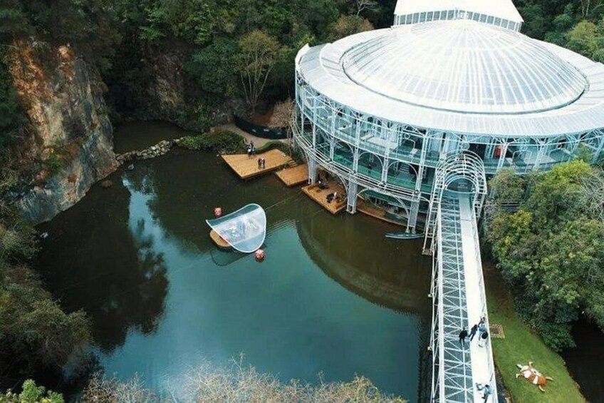 A glasshouse structure with a circular roof, a wooden dock, and a small boat in a body of water surrounded by greenery. 2