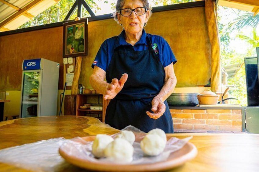 An elderly person in a blue shirt and apron, gesturing in front of a table with a plate of dumplings. 5