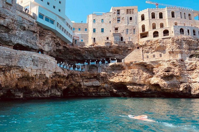Ein Schwimmer im klaren, blauen Meer, Felsenküste und historische Gebäude. 2