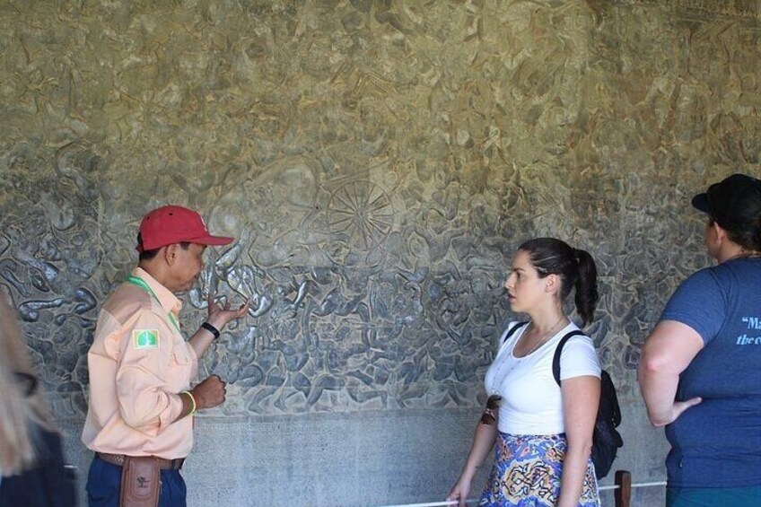 Un mur à la texture particulière, orné de sculptures complexes, une personne portant une veste rose et une casquette rouge, et une autre vêtue d’un haut blanc et d’une jupe à motifs. 2