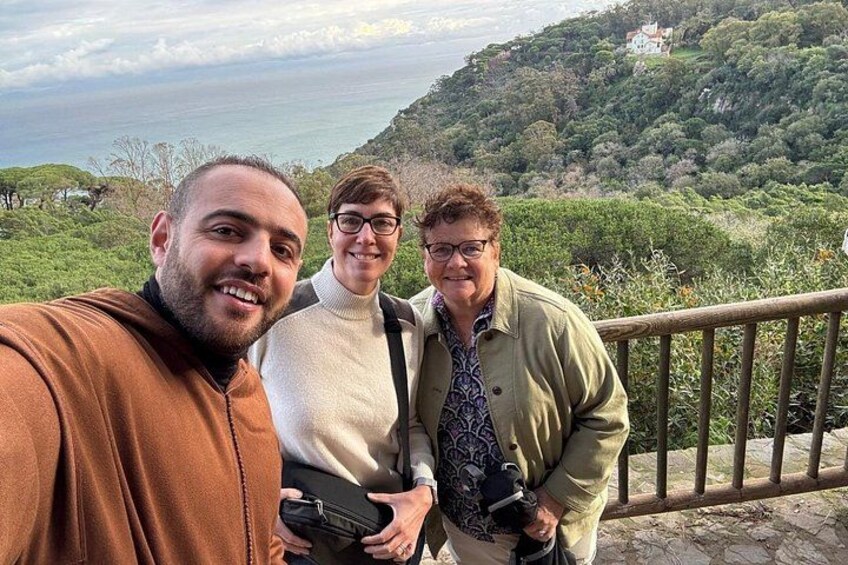 Tres personas de pie en una barandilla de madera con vistas a un paisaje montañoso, vegetación y el mar al fondo. 2