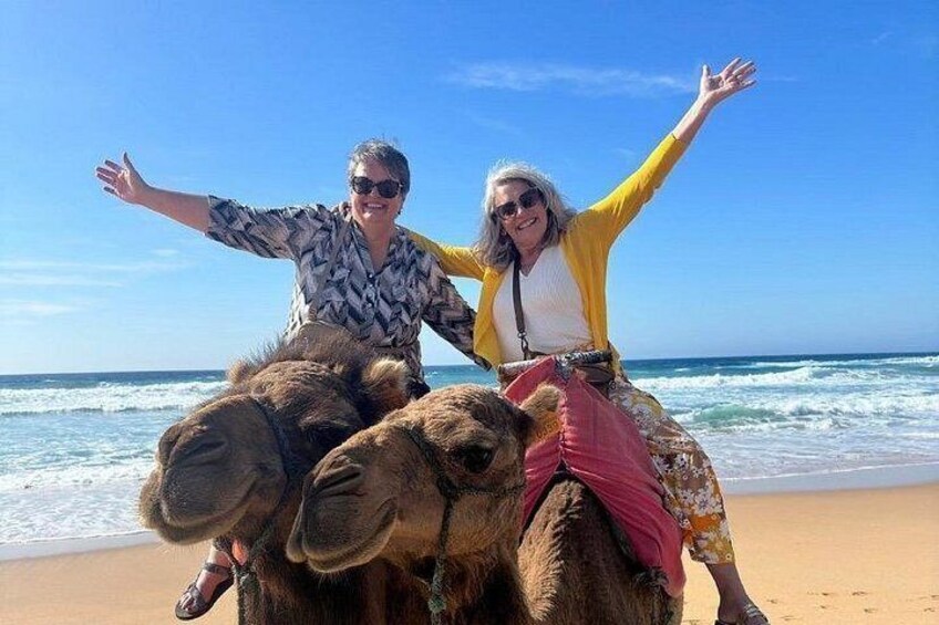 Dos personas montando camellos en una playa, con el océano al fondo. 1