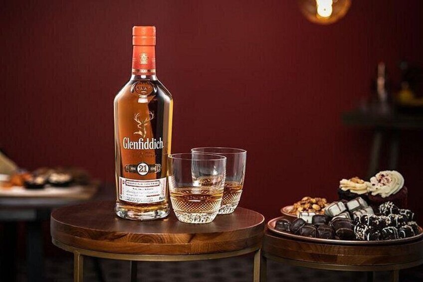 Une bouteille de whisky Glenfiddich, deux verres et une assiette remplie de diverses collations sur une table en bois. 1